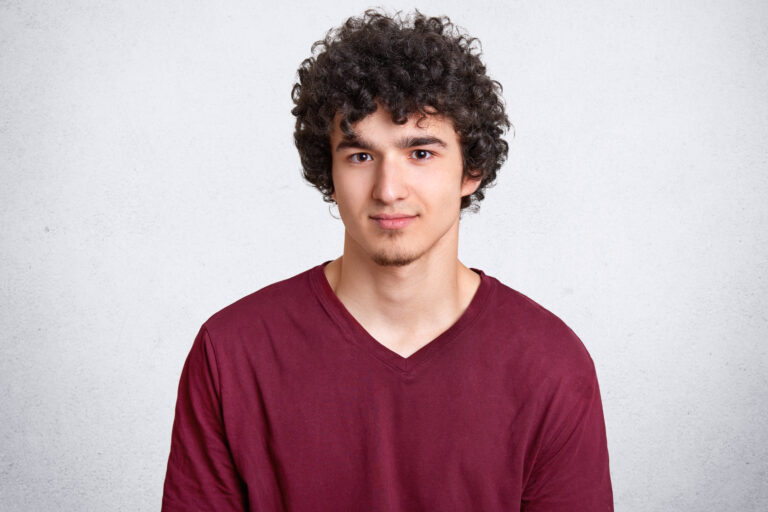 Headshot of attractive curly youngster looks seriously at camera, has appealing look, spends free time with friends, dressed in casual outfit, isolated over white concrete wall